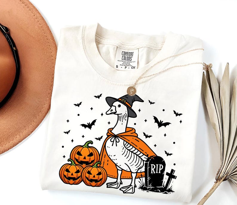 Puede incluir: Una camiseta blanca con una ilustraci&oacute;n en blanco y negro de un ganso con un sombrero de bruja y una capa. El ganso est&aacute; de pie junto a una l&aacute;pida que dice "RIP". Tambi&eacute;n hay calabazas y murci&eacute;lagos en la ilustraci&oacute;n.