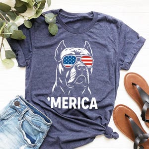 May include: Navy blue t-shirt with a white graphic of a dog wearing sunglasses in the style of the American flag. The word "'MERICA" is printed below the dog. The shirt is paired with blue jeans and brown sandals.