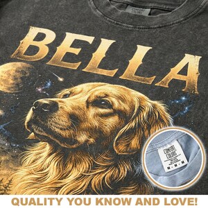 May include: Dark gray t-shirt with the name "BELLA" in gold lettering above a detailed illustration of a golden retriever dog. The design includes a space-themed background with planets and stars. The shirt has a "COMFORT COLORS" tag.