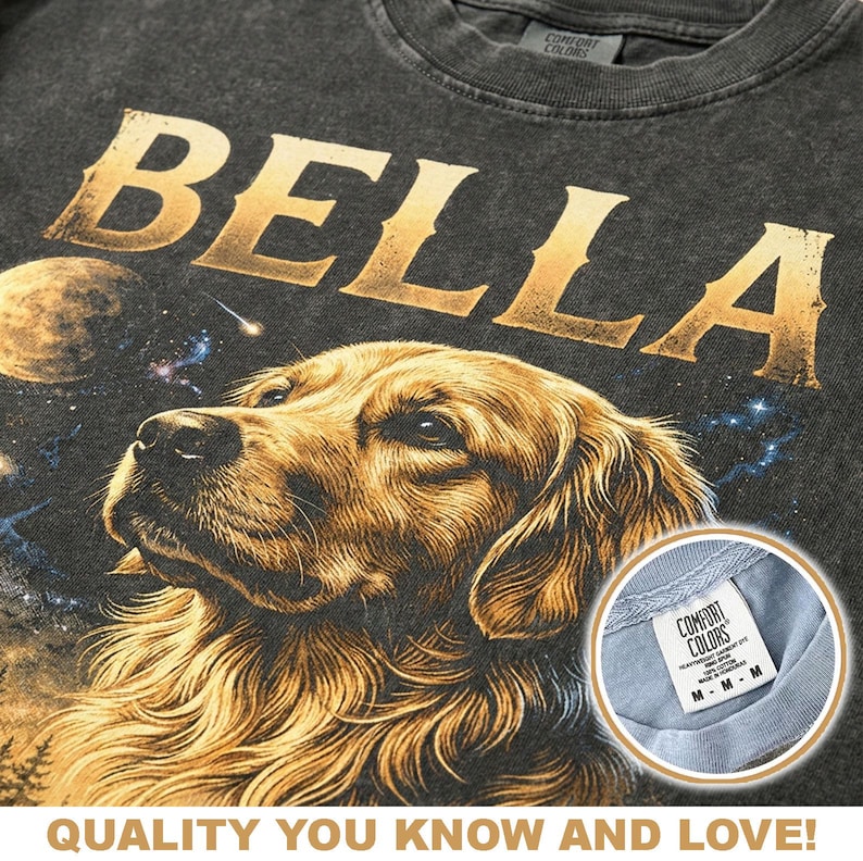 May include: Dark gray t-shirt with the name "BELLA" in gold lettering above a detailed illustration of a golden retriever dog. The design includes a space-themed background with planets and stars. The shirt has a "COMFORT COLORS" tag.