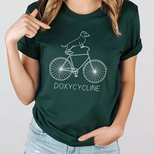 May include: A dark green t-shirt with a white outline of a dachshund riding a bicycle. The text "DOXYCYCLINE" is printed below the bicycle.