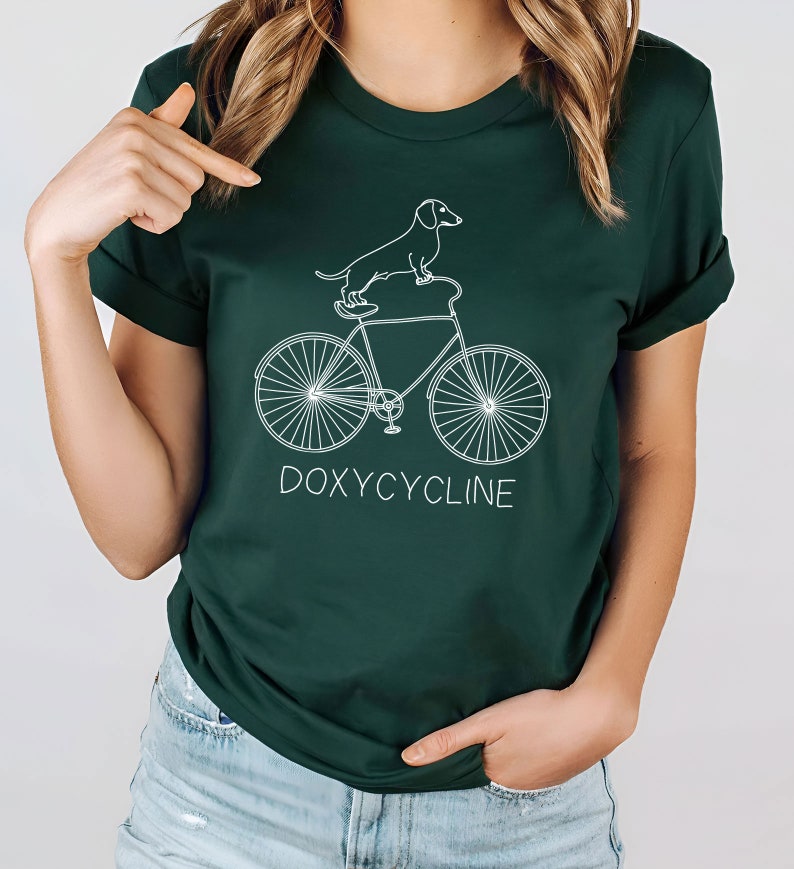 May include: A dark green t-shirt with a white outline of a dachshund riding a bicycle. The text "DOXYCYCLINE" is printed below the bicycle.