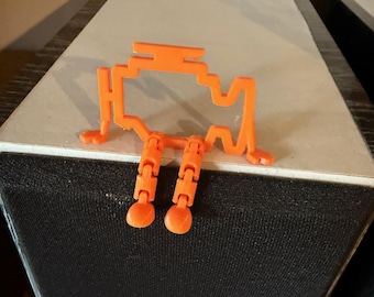 May include: An orange 3D printed figurine of a car engine with legs. The figurine is sitting on top of a black speaker.
