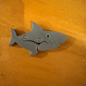 May include: A gray shark-shaped wooden puzzle with articulated joints. The shark has fins, a tail, and a detailed mouth. The puzzle is designed to be assembled and disassembled, offering an engaging activity for children and adults.