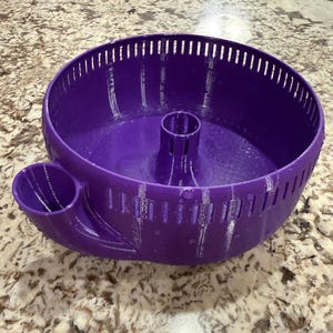 May include: A purple, circular pet accessory with a central cylinder and an attached side compartment. The item has a slotted design around the rim and is made of a solid, smooth material. The accessory sits on a speckled surface.