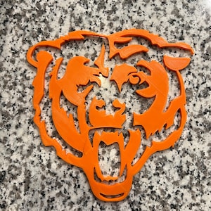 Peut inclure: Silhouette de tête d'ours orange imprimée en 3D. L'ours a la gueule ouverte avec des dents acérées et une expression féroce.