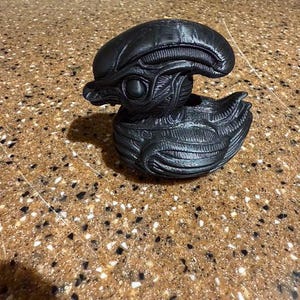 May include: A black, sculpted figurine of an alien head, featuring detailed textures and a smooth, glossy finish. The design is inspired by the iconic creature from the Alien film franchise. The figurine is a decorative item.