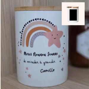 Peut inclure: Un bougeoir en céramique blanc avec un couvercle en bois. Le design présente un arc-en-ciel pastel, une étoile souriante et le texte "Merci Nounou Jeanne de m'aider à grandir Camille."