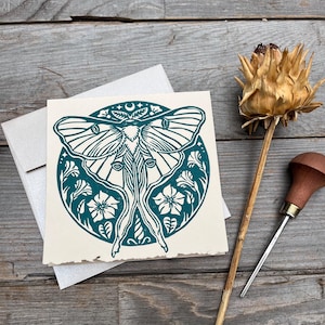 May include: A teal and cream-coloured greeting card with a detailed moth design within a circular floral frame. The card is with a silver envelope, a dried thistle, and a wood carving tool, all on a weathered wooden surface.