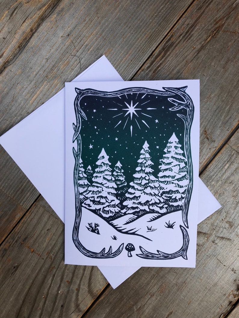 Puede incluir: Una ilustraci&oacute;n en blanco y negro de una escena de bosque nevado con una gran estrella en el cielo nocturno. La ilustraci&oacute;n est&aacute; enmarcada por un borde decorativo.