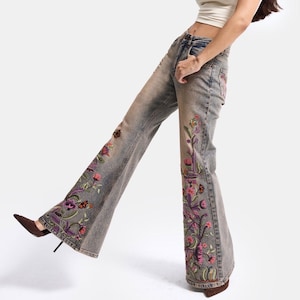 Op de afbeelding: Een uitlopende spijkerbroek van lichtgewassen denim met ingewikkeld, veelkleurig bloemenborduursel langs de buitenkant van de pijpen, gecombineerd met een crèmekleurige, nauwsluitende crop top met korte mouwen en donkerbruine pumps.