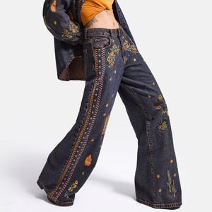 May include: A dark wash denim jacket and matching wide-leg jeans set featuring intricate embroidery of flowers, butterflies, cacti, and small bottles. The denim fabric has a deep charcoal tone with decorative vertical side stripes and star accents.