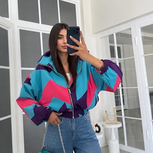 Può includere: Una giacca con zip caratterizzata da un motivo geometrico nei colori turchese brillante, rosa acceso e viola scuro, abbinata a jeans blu e a una piccola borsa color verde acqua con tracolla a catena argentata. La giacca presenta collo alto e polsini elasticizzati.
