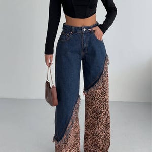 May include: Denim jeans with a dark wash and a distinctive leopard print panel on the lower leg. The jeans have a high waist and a frayed edge detail. The outfit includes a black long-sleeved crop top.