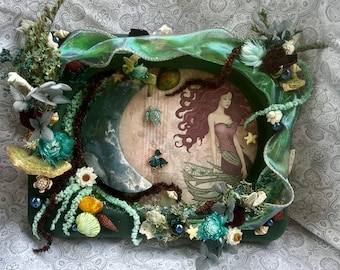 May include: A rectangular green frame decorated with dried flowers, seashells, and a crescent moon shape. Inside, an illustration of a long-haired figure in a dress is displayed alongside small turtle and fish charms on a light background.