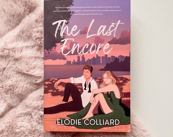 May include: A book titled "The Last Encore" by Elodie Colliard. The cover features a couple sitting in front of a city skyline at sunset. The book is resting on a fluffy, light pink surface.