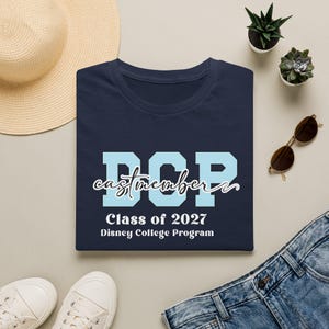 May include: A navy blue t-shirt featuring the light blue letters DCP, the word castmember in white cursive script, and the text Class of 2027 Disney College Program printed in white on the front of the garment.