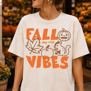 May include: An ivory short-sleeve t-shirt featuring orange graphic text that reads FALL VIBES with the words stay cozy in smaller script. The design includes a pumpkin, a maple leaf, an apple, and a squirrel illustration in orange ink.