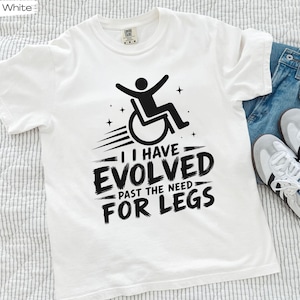 May include: A white short-sleeve t-shirt featuring a black graphic of a person in a wheelchair with motion lines and stars. The text reads: I HAVE EVOLVED PAST THE NEED FOR LEGS. The shirt has a crew neckline and a visible brand tag at the inner collar.