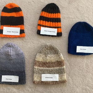 May include: Five knitted beanies in various patterns and colors. One orange and black striped, one black and orange striped, one solid blue, one grey striped, and one brown striped. Each beanie features a white label with its color description.