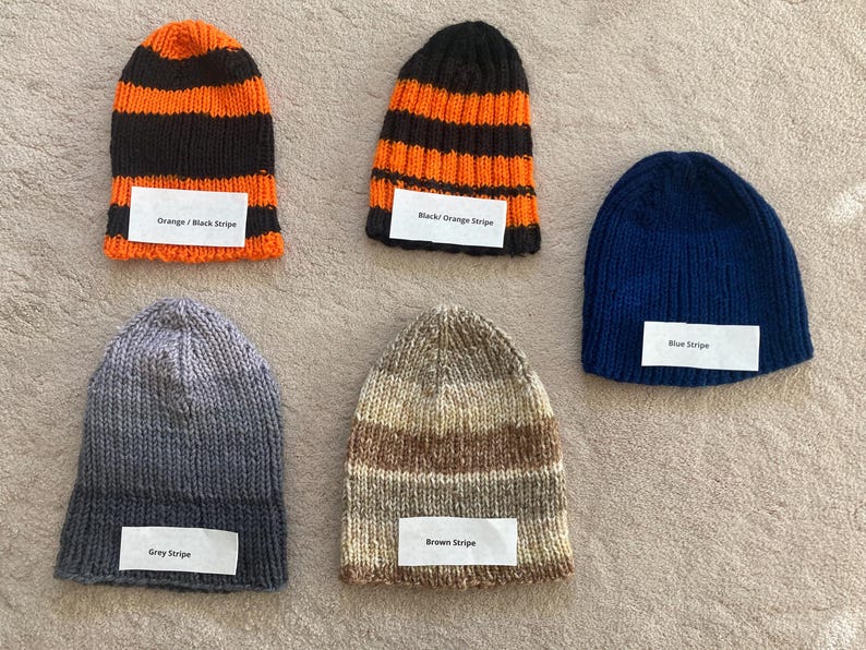 May include: Five knitted beanies in various patterns and colors. One orange and black striped, one black and orange striped, one solid blue, one grey striped, and one brown striped. Each beanie features a white label with its color description.