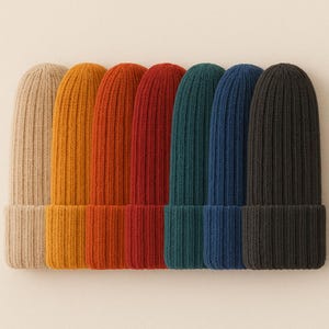 May include: A collection of seven ribbed knit beanies arranged in a row, featuring a spectrum of colors including beige, mustard yellow, burnt orange, red, teal, navy blue, and black. Each beanie has a wide folded cuff and a vertical knit pattern.