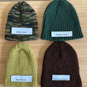 May include: Four knitted beanies in different colors. The top left is green camo, the top right is hunter green, the bottom left is apple green, and the bottom right is chocolate brown. Each beanie has a white label with its color name printed on it.
