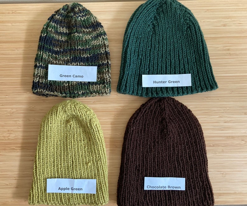 May include: Four knitted beanies in different colors. The top left is green camo, the top right is hunter green, the bottom left is apple green, and the bottom right is chocolate brown. Each beanie has a white label with its color name printed on it.