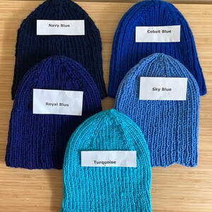 May include: Five knitted beanies in various shades of blue, each with a white label. The labels read Navy Blue, Cobalt Blue, Royal Blue, Sky Blue, and Turquoise. These hats feature a ribbed texture and are arranged in a cluster on a flat surface.