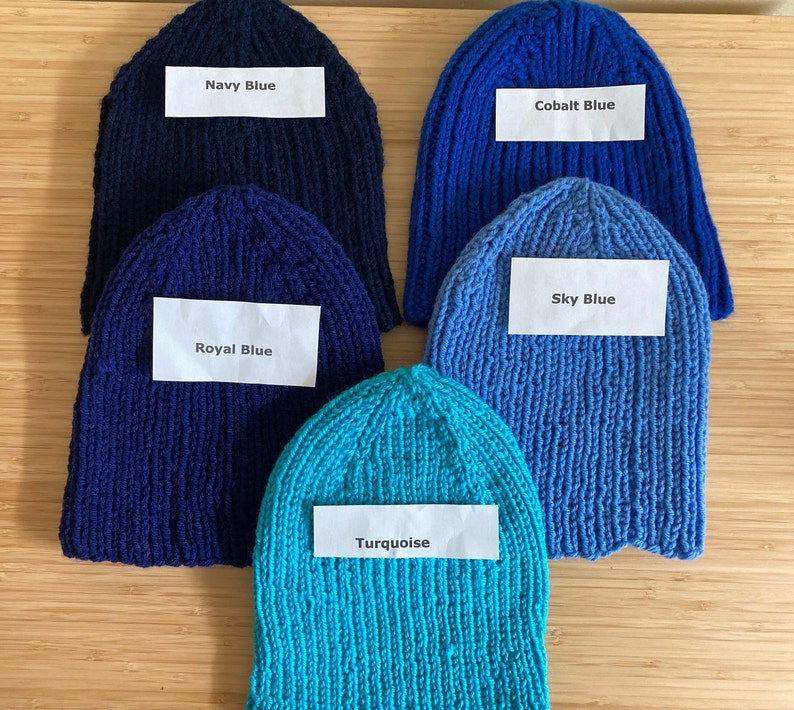 May include: Five knitted beanies in various shades of blue, each with a white label. The labels read Navy Blue, Cobalt Blue, Royal Blue, Sky Blue, and Turquoise. These hats feature a ribbed texture and are arranged in a cluster on a flat surface.