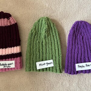 May include: Three knit beanies: one with dark maroon and light pink stripes labeled Bubblegum Stripe, one solid mint green labeled Mint green, and one solid purple labeled Purple Rain. Each hat features a vertical ribbed texture pattern.