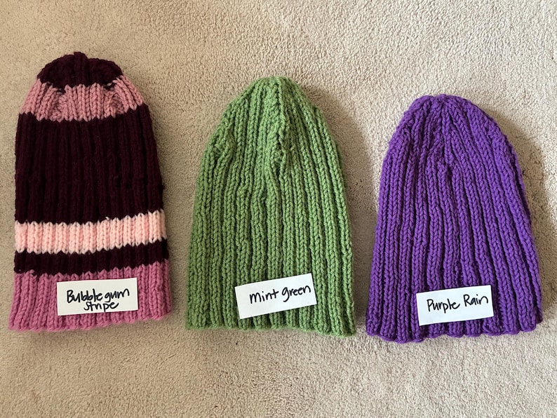 May include: Three knit beanies: one with dark maroon and light pink stripes labeled Bubblegum Stripe, one solid mint green labeled Mint green, and one solid purple labeled Purple Rain. Each hat features a vertical ribbed texture pattern.