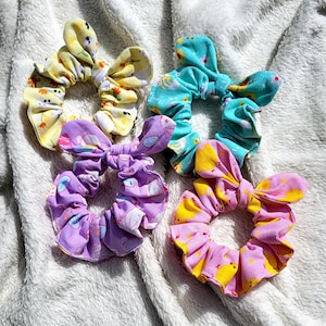 May include: Four colourful hair scrunchies with different patterns. The scrunchies are made of fabric and have a bow on top. The patterns include yellow chicks on pink, purple bunnies on lavender, blue flowers on turquoise, and yellow chicks on yellow.