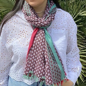 Peut inclure: Une écharpe à motifs marrons, verts et rouges avec des motifs floraux blancs. L'écharpe est nouée autour du cou et a des franges aux extrémités.