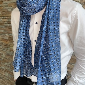 Può includere: Sciarpa blu con un motivo geometrico di quadrati neri e bianchi, drappeggiata su una camicia bianca con colletto. La sciarpa è realizzata in materiale leggero ed è indossata da una persona che indossa pantaloni neri.