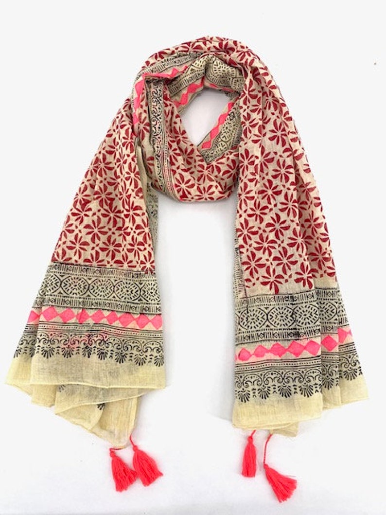 May include: A cream-colored cotton scarf with a red and black floral print and pink accents. The scarf has a black and white border with a geometric pattern and tassels on the ends.