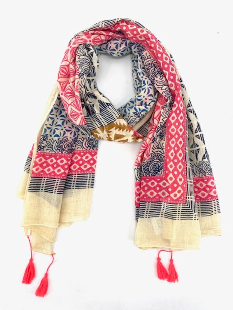 May include: A cream-colored cotton scarf with a pink, blue, and brown geometric pattern. The scarf has tassels on each end.