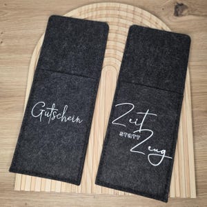 K&ouml;nnte beinhalten: Zwei dunkelgraue Filzh&uuml;llen mit wei&szlig;er Schrift. Auf einer steht "Gutschein" und auf der anderen "Zeit statt Zeug". Die H&uuml;llen liegen auf einem Holztablett mit geschwungenem Design.