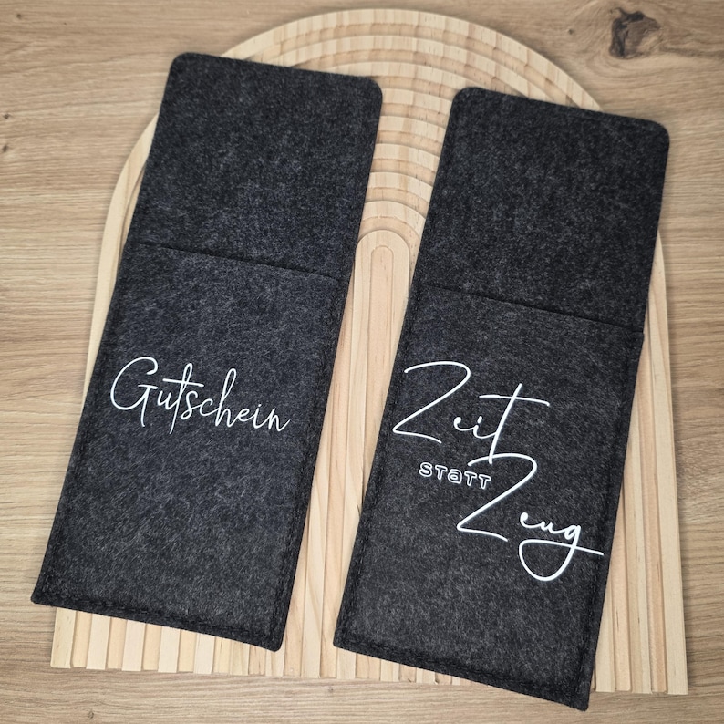 K&ouml;nnte beinhalten: Zwei dunkelgraue Filzh&uuml;llen mit wei&szlig;er Schrift. Auf einer steht "Gutschein" und auf der anderen "Zeit statt Zeug". Die H&uuml;llen liegen auf einem Holztablett mit geschwungenem Design.