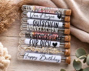 Könnte beinhalten: Eine Sammlung von Glasröhrchen mit Korken, die jeweils dekorative Elemente und ein bedrucktes Etikett enthalten. Die Etiketten tragen Aufschriften wie "Happy Birthday" und "Für dich". Die Röhrchen sind auf einer hellen Holzoberfläche angeordnet, mit einem weichen, neutralfarbenen Stoff im Hintergrund.