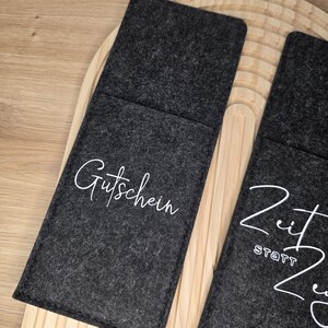 K&ouml;nnte beinhalten: Dunkelgraue Filz-Geschenkkartenhalter mit wei&szlig;er Schrift. Ein Halter tr&auml;gt die Aufschrift "Gutschein" und der andere "Zeit statt Zeug". Die Halter liegen auf einer hellen Holzoberfl&auml;che.