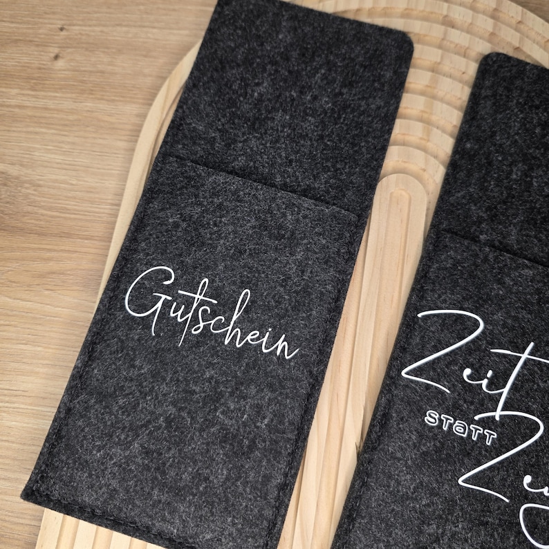 K&ouml;nnte beinhalten: Dunkelgraue Filz-Geschenkkartenhalter mit wei&szlig;er Schrift. Ein Halter tr&auml;gt die Aufschrift "Gutschein" und der andere "Zeit statt Zeug". Die Halter liegen auf einer hellen Holzoberfl&auml;che.