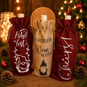 Könnte beinhalten: Drei Weinflaschen-Geschenktaschen aus Jute. Zwei Taschen sind dunkelrot mit weißem Text "Frohes Fest" und "Cheers!" sowie einem Wichtel-Motiv. Die mittlere Tasche ist naturfarben mit schwarzem Text "FRÖHLICHE WEIN ACHTEN" und einem Wichtel.