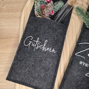 K&ouml;nnte beinhalten: Dunkelgrauer Filz-Geschenkkartenhalter mit dem Wort "Gutschein" in wei&szlig;er Schrift. Der Halter enth&auml;lt ein Glasfl&auml;schchen mit Korken und ist mit k&uuml;nstlichen Beeren und Tannennadeln verziert. Der Halter befindet sich auf einer Holzoberfl&auml;che.