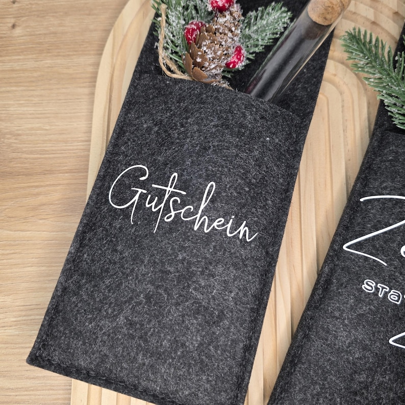 K&ouml;nnte beinhalten: Dunkelgrauer Filz-Geschenkkartenhalter mit dem Wort "Gutschein" in wei&szlig;er Schrift. Der Halter enth&auml;lt ein Glasfl&auml;schchen mit Korken und ist mit k&uuml;nstlichen Beeren und Tannennadeln verziert. Der Halter befindet sich auf einer Holzoberfl&auml;che.