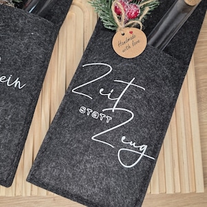 K&ouml;nnte beinhalten: Dunkelgrauer Filz-Weinflaschenhalter mit wei&szlig;er Schrift "Zeit statt Zeug". Ein kleines Etikett lautet "Handmade with love". Der Halter ist mit einem Zweig aus Gr&uuml;n und roten Beeren verziert. Eine Glasflasche ist sichtbar.