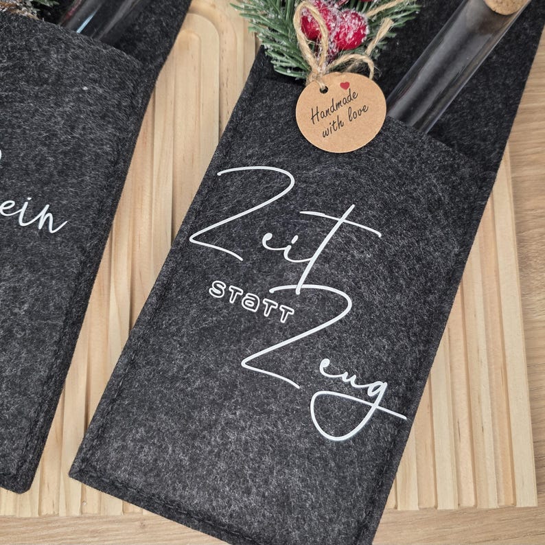 K&ouml;nnte beinhalten: Dunkelgrauer Filz-Weinflaschenhalter mit wei&szlig;er Schrift "Zeit statt Zeug". Ein kleines Etikett lautet "Handmade with love". Der Halter ist mit einem Zweig aus Gr&uuml;n und roten Beeren verziert. Eine Glasflasche ist sichtbar.