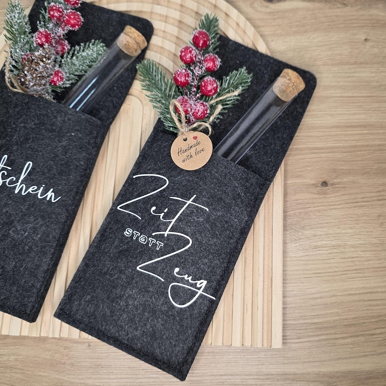 K&ouml;nnte beinhalten: Zwei dunkelgraue Filztaschen, jede mit einem Glasr&ouml;hrchen mit Korken. Eine Tasche hat das Wort "Gutschein" in wei&szlig;er Schrift, die andere "Zeit Statt Zeug". Beide sind mit k&uuml;nstlichen Beeren und Gr&uuml;nzeug verziert. Ein kleines Etikett lautet "Handmade with love".