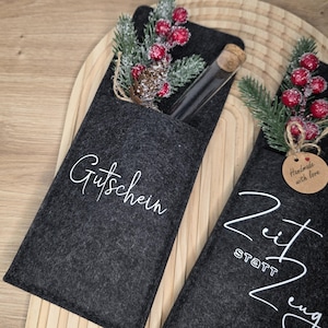 K&ouml;nnte beinhalten: Zwei dunkelgraue Filz-Geschenkkartenhalter mit wei&szlig;er Schrift. Einer tr&auml;gt die Aufschrift "Gutschein" und der andere "Zeit statt Zeug". Jeder Halter hat ein kleines Glasr&ouml;hrchen und ist mit k&uuml;nstlichen Beeren, Tannennadeln und einem Tannenzapfen verziert.
