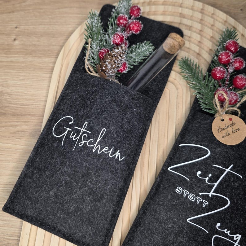 K&ouml;nnte beinhalten: Zwei dunkelgraue Filz-Geschenkkartenhalter mit wei&szlig;er Schrift. Einer tr&auml;gt die Aufschrift "Gutschein" und der andere "Zeit statt Zeug". Jeder Halter hat ein kleines Glasr&ouml;hrchen und ist mit k&uuml;nstlichen Beeren, Tannennadeln und einem Tannenzapfen verziert.